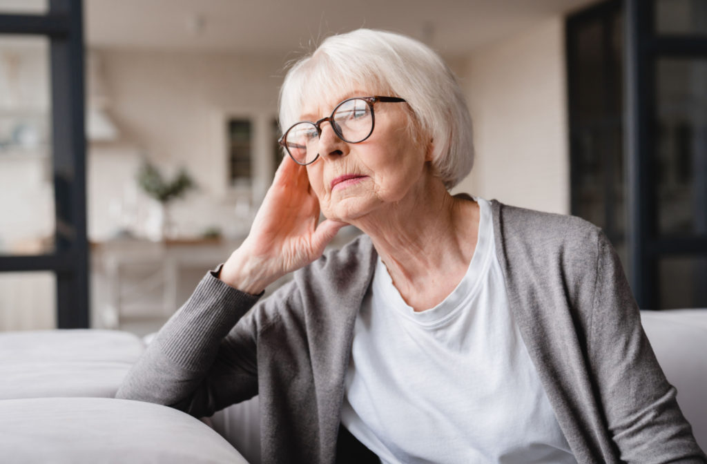 A senior woman with glasses sitting on a couch appears to be sad and looking outside the window.