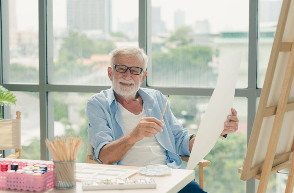A smiling senior man sitting in an art studio and holding a paintbrush in one hand and a piece of paper in the other hand.