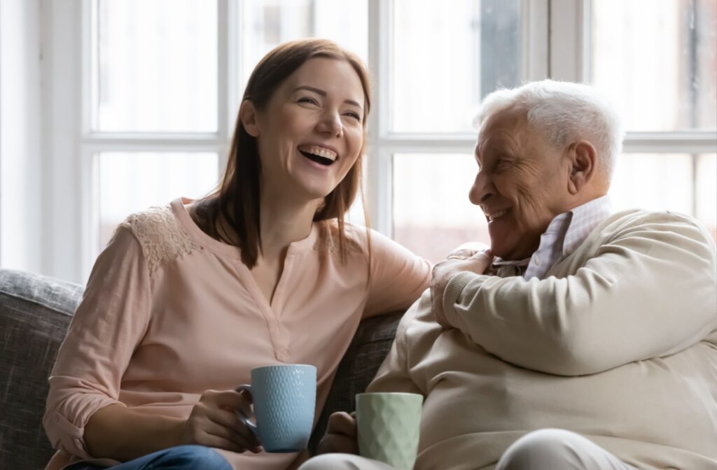 Adult child and senior parent drinking tea while talking and laughing on a cozy couch.
